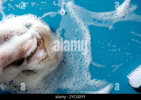 Le mani tengono una spugna per la pulizia e puliscono una schiuma saponata su una piastrella blu. sfondo blu. Concetto di pulizia, servizio di pulizia. Banner. Disposizione piatta, vista dall'alto Foto Stock