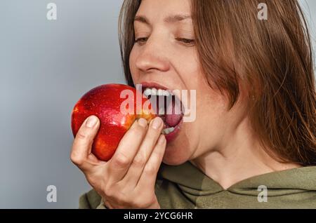 La donna morde la mela rossa, mostrando i denti. Capelli castani. Apple è tenuto in mano destra su sfondo grigio Foto Stock