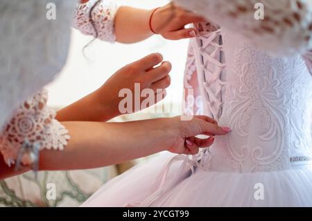 Damigella aiutando slanciata allacciatura sposa il suo matrimonio abito bianco, abbottonatura sul delicato merletto pattern con mantello soffice sulla vita. Mattina prepara nuziale Foto Stock