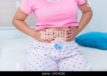 Una donna tocca il suo addome seduto sul letto; i crampi mestruali sono comuni ogni mese nelle donne in età fertile. Primo piano. Foto Stock