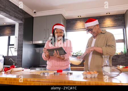 Periodo natalizio, coppia senior multirazziale che indossa cappelli di babbo natale da cuocere, usando un tablet, a casa Foto Stock