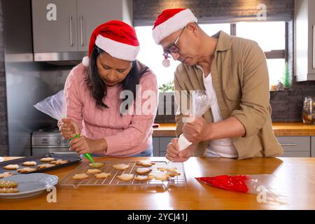 Coppia senior multirazziale che decora biscotti di natale, indossa cappelli di babbo natale, a casa Foto Stock