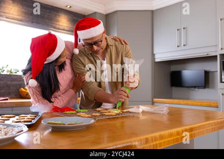 Coppia senior multirazziale che decora biscotti di natale, indossa cappelli di babbo natale, a casa Foto Stock