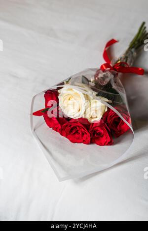 Un bellissimo bouquet di rose rosse e bianche avvolte in plastica trasparente, posto su un letto ben fatto con morbide lenzuola bianche e un tocco decorativo di pillo Foto Stock