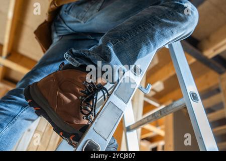 Un lavoratore è posizionato con cura su una scala, indossando stivali robusti e jeans denim, impegnato in lavori di ristrutturazione domestica sotto le luci luminose del soffitto. Foto Stock