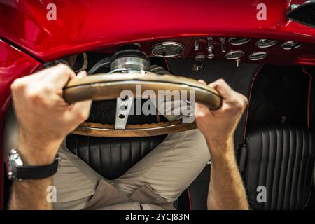 Una persona afferra il volante in legno di una classica auto rossa, concentrandosi sulla guida su una strada aperta sotto la luce del sole. Foto Stock