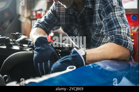 Una persona si concentra sulla riparazione del motore di un'auto in un garage, indossando guanti blu e una camicia a quadri, dimostrando dedizione e abilità nel lavoro automobilistico. Foto Stock