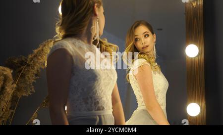 La sposa si ammira con un abito in pizzo bianco mentre posa di fronte a uno specchio Foto Stock