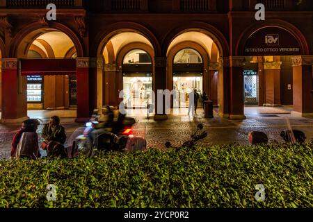Gelateria in serata dietro Portici illuminati e passanti con la loro Vespa. Piazza Cavour, Bologna, Emilia-Romagna, Italia Foto Stock