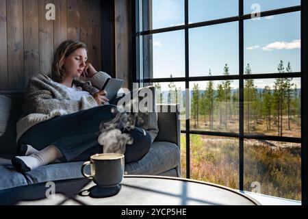 Donna seduta su un comodo divano, che legge un libro vicino a una grande finestra in una cabina con una bevanda calda accanto a lei, godendo la vista della foresta all'esterno Foto Stock