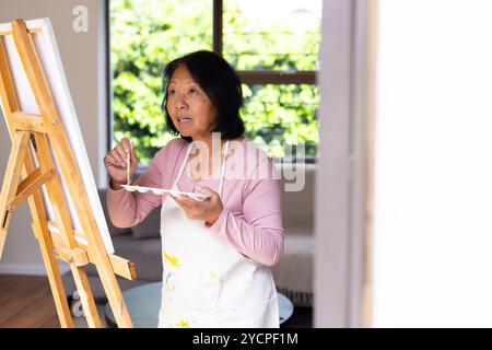 donna asiatica senior che dipinge su tela a casa, concentrata e creativa Foto Stock