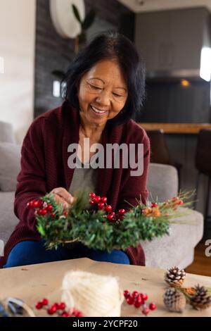 Donna asiatica sorridente che crea la corona di Natale a casa, godendo dei preparativi per le vacanze Foto Stock