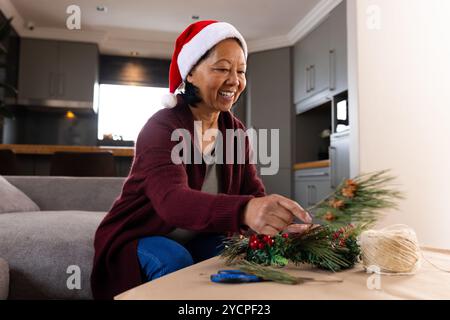 Creazione di corona di Natale, donna asiatica anziana sorridente durante i preparativi per le vacanze a casa Foto Stock