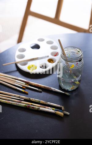 Forniture artistiche sul tavolo con pennelli, tavolozza e vasetto d'acqua, a casa Foto Stock