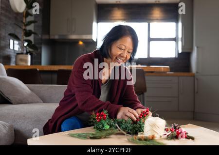 Natale, donna asiatica sorridente che crea la corona di natale a casa Foto Stock