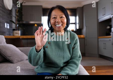 Durante una videochiamata, una donna asiatica anziana sorridente che sventola a casa, godendo un caldo momento natalizio Foto Stock