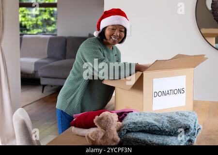 Donna asiatica sorridente nel cappello di Babbo Natale che prepara una scatola di donazioni a casa durante il Natale Foto Stock