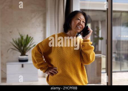 Donna asiatica anziana in maglione giallo che parla su smartphone a casa, sorride calorosamente Foto Stock