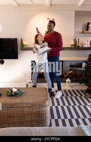 Coppia multirazziale che ride e balla accanto al caminetto, festeggiando il Natale a casa Foto Stock