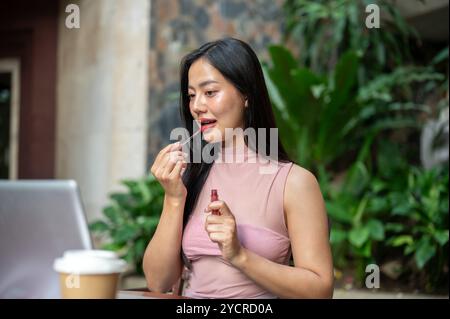 Una bella donna asiatica che applica il rossetto mentre lavora al suo laptop, siede a un tavolo all'aperto, lavora in remoto in un bar. Foto Stock