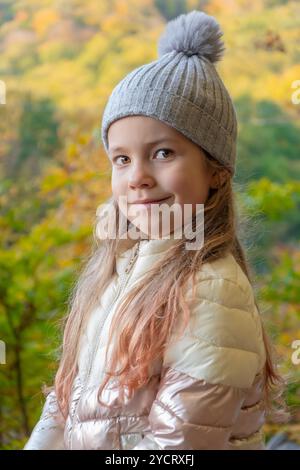 Una ragazza allegra si trova in una foresta luminosa piena di colori autunnali, indossando un cappello grigio a maglia e una giacca bianca. La luce del sole penetra attraverso la t Foto Stock