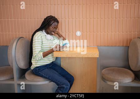 Una giovane donna utilizza felicemente il suo smartphone in un accogliente bar, incarnando lo spirito spensierato dei millennial e della gen z Foto Stock