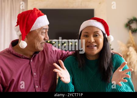 Durante una videochiamata, una coppia senior multirazziale sorridente che indossa cappelli di babbo natale per celebrare il natale a casa Foto Stock