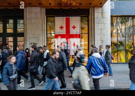 Tbilisi, Georgia. 23 ottobre 2024. Una folla di persone passa davanti a una grande bandiera georgiana esposta su un edificio nel centro di Tbilisi mentre la città ospita una manifestazione politica. Il movimento per le strade riflette l'urgenza e l'attesa che portano alle elezioni. Catturato durante la campagna pre-elettorale del Partito del sogno georgiano a Tbilisi, Georgia, mercoledì 23 ottobre 2024. (VX Photo/ Vudi Xhymshiti) crediti: VX Pictures/Alamy Live News Foto Stock