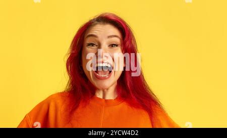 Ritratto di una giovane donna rossa super eccitata con abiti arancioni, che esprime emozionalmente emozioni e vittoria su sfondo giallo brillante. Foto Stock