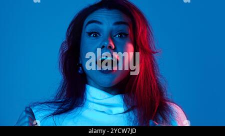 Ritratto di una giovane donna super eccitata che esprime emozionalmente eccitazione e vittoria su sfondo blu in studio sotto la luce del neon. Foto Stock