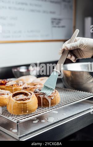 Un pasticcere vegano decora meticolosamente un lotto di dolci alla cannella appena sfornati con glassa in una panetteria insieme a diverse ciambelle smaltate, mostrando il cu Foto Stock