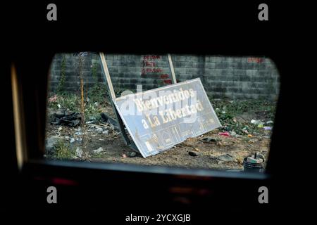 Un cartello intempestivo e abbandonato che legge Bienvenidos a la Libertad giace tra la spazzatura sparsa da un muro di El Salvador, visto attraverso un'auto Foto Stock