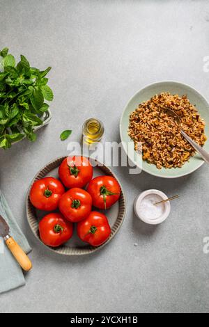 Vista dall'alto dei pomodori rossi vivaci in un recipiente di ceramica accanto a un piatto di riso integrale stagionato con noci ed erbe aromatiche, accompagnato da menta fresca, olio d'oliva, a. Foto Stock