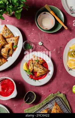 Deliziosi samosas al forno croccanti su sfondo rosso, guarniti con menta fresca e semi di sesamo, accompagnati da una vivace salsa di pomodoro che ritrae una verga Foto Stock