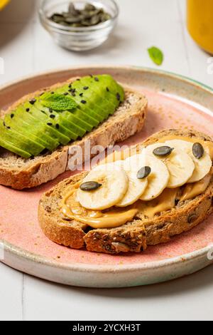 Immagine elegante con toast all'avocado e toast al burro di arachidi con fette di banana, guarnite con semi, su un piatto rosa tenue sfondo chiaro aggiunge un rif Foto Stock