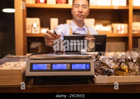 Il dipendente asiatico di una cioccolateria seleziona e pesa una varietà di cioccolatini su scala digitale al banco cassa, fornendo servizio in un ambiente accogliente Foto Stock