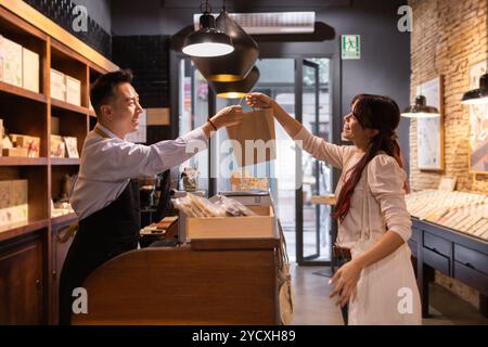 Un uomo asiatico consegna un sacchetto di carta a una donna sorridente in una pittoresca cioccolateria, mostrando un eccellente servizio clienti e un invitante interio illuminato calorosamente Foto Stock