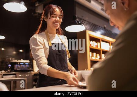 Una gioiosa impiegata del negozio serve una barretta di cioccolato a un cliente presso un negozio di cioccolato ben illuminato, l'illuminazione ambientale e gli scaffali dei cioccolatini nella ba Foto Stock