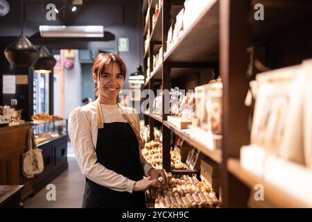 Un allegro dipendente in un negozio di cioccolato organizza vari dolci su scaffali in legno, mostrando una vasta selezione di cioccolatini gourmet in un mese Foto Stock