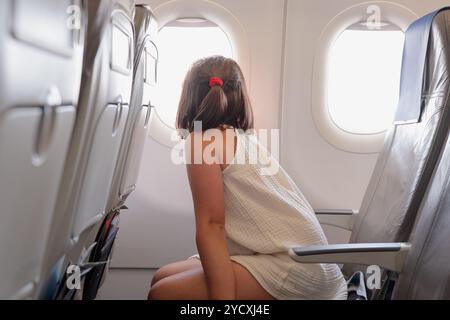 Vista posteriore di una ragazza irriconoscibile seduta all'interno di un aereo, che guarda fuori dalla finestra, vestita con un leggero abito estivo con i capelli legati in una patta Foto Stock