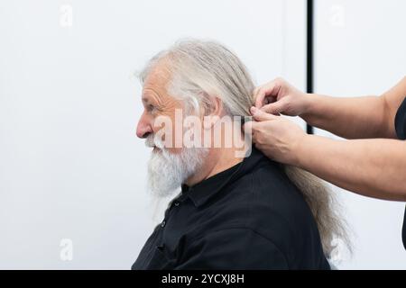 Un uomo anziano con una lunga barba grigia sorride mentre una donna raccoglie i suoi lunghi capelli grigi, preparandoli per la donazione Foto Stock