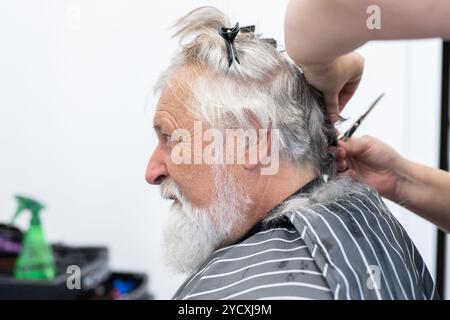 Un uomo anziano partecipa a un evento di donazione dei capelli, ottenendo i suoi lunghi capelli grigi tagliati da un parrucchiere in un ambiente professionale Foto Stock