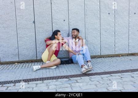 La coppia gay condivide un momento di gioia seduto contro un muro in una giornata di sole, che riflette le diverse culture dell'America Latina e dell'afroamericano heritag Foto Stock