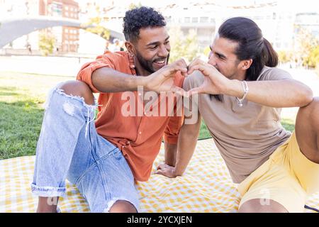Una coppia gay affettuosa forma il cuore con le mani mentre si rilassa su una coperta da picnic in un vivace parco cittadino, riflettendo la diversità e la diversità Foto Stock