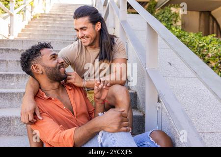 Momento di gioia tra una coppia gay mentre si siedono sulle scale della città, mostrando la loro relazione amorosa entrambi uomini, un afroamericano e uno ispanico, e. Foto Stock