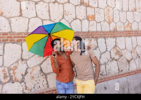 Le coppie gay godono di una giornata di sole in città, riparate sotto un ombrello colorato, a simboleggiare l'orgoglio LGBTQIA+ sorridente e coinvolgente affettuosamente, t Foto Stock