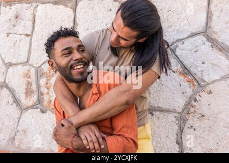 Un momento gioioso catturato tra due uomini gay, uno che abbraccia l'altro contro un muro di pietra testurizzato in un ambiente urbano, mettendo in risalto il loro multietnio Foto Stock