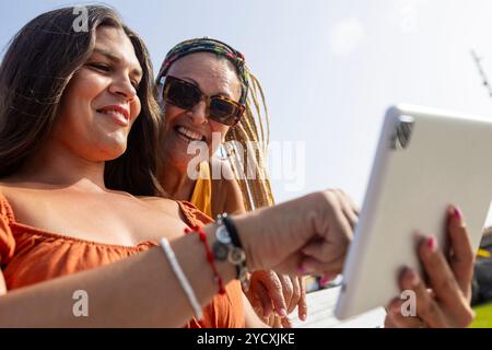 Una madre sorridente e la figlia transgender si divertono insieme all'aperto guardando un tablet digitale. Foto Stock