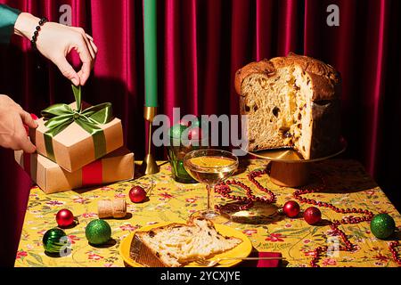 Le mani di una donna posizionano i regali di Natale avvolti su un tavolo festivo con panettone, champagne, candele e colorate decorazioni natalizie Foto Stock