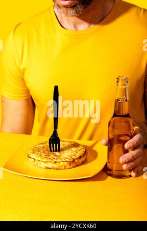 Uomo che indossa una T-shirt gialla, pronto a mangiare una tortilla spagnola da un piatto giallo, mentre tiene una bottiglia di birra chiara lo sfondo e il tavolo sono u Foto Stock